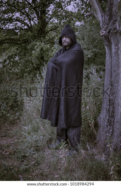 Hooded man wearing a cloak in a mysterious\
fantasy forest setting.