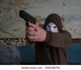 Hooded Robber With A Gun On Old Tiled Wall Background. Man Wear White Hockey Mask And Warm Pullover. 