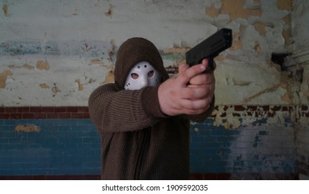 Hooded Robber With A Gun On Old Tiled Wall Background. Man Wear White Hockey Mask And Warm Pullover. 