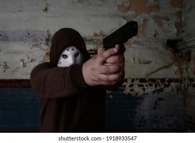 Hooded Robber With A Gun On Old Tiled Wall Background. Man Wear White Hockey Mask And Warm Pullover. 