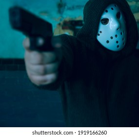 Hooded Robber With A Gun On Old Tiled Wall Background. Man Wear White Hockey Mask And Warm Pullover. 