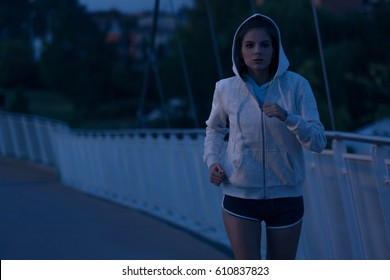 Hooded young woman running by night in the park. Fitness and workout wellness concept. Sports outdoors. Cityscape Background - Powered by Shutterstock