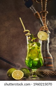 Hookah (shisha) and glass of Mojito cocktail with lime and ice on dark brown wooden table on dark background with smoke. Weekend or holiday party