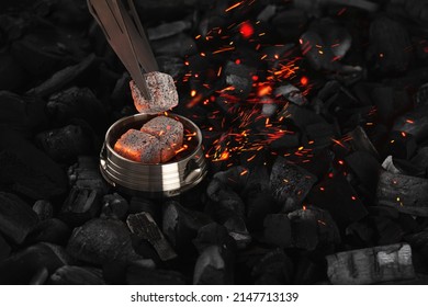 With Hookah Tongs, They Shift And Blow On Burning Cubes Of Coconut Charcoal In A Metal Kaloud, Sparks Fly On A Black Background. Horizontal Orientation, Close-up, Focus In The Foreground, No People.