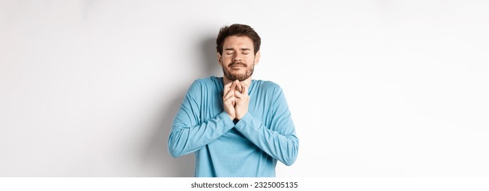 Hopeful caucasian guy in sweatshirt cross fingers for good luck. Young man making wish, praying with eyes closed and dreamy smile, standing over white background.