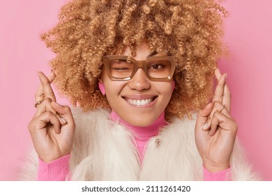 Hopeful Cheerful Woman Bites Lips Keeps Fingers Crossed Makes Wish Hopes Dreams Come True Wears Winter Fur Coat Spectacles Poses Against Pink Background Prays For Good Results. Body Language