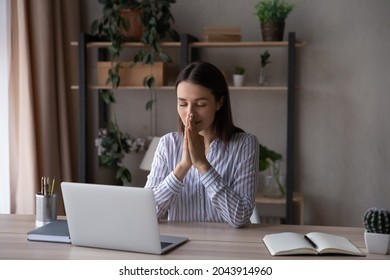 Hopeful religious businesswoman joining hands in prayer, sitting at home office desk with laptop, wishful mindful young woman with closed eyes dreaming, asking help, begging for good work result