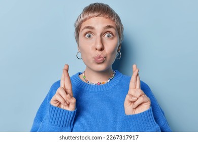 Hopeful short haired woman keeps lips rounded crosses fingers believes in good luck wears casual jumper earrings and necklace isolated over blue background wishes for good luck prays for something - Shutterstock ID 2267317917