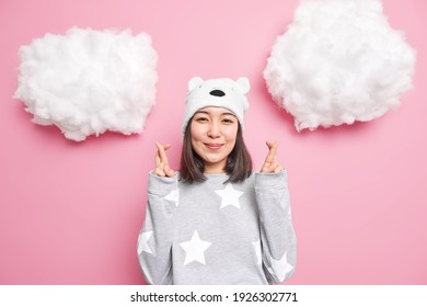 Hopeful smiling gentle Asia woman wears pajama bear hat crosses fingers waits for good news makes wish or prays anticipates something happen isolated over pink background with white clouds above