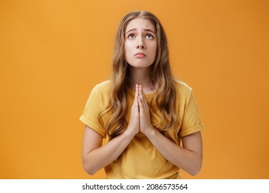 Hopeful uneasy and worried gloomy faithful girl in t-shirt holding hands in pray against chest looking up with sad look praying making wish for good well of family posing over orange wall - Shutterstock ID 2086573564