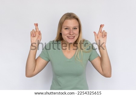 Hopeful young woman crossing fingers. Portrait of happy Caucasian female model with fair hair in green T-shirt looking at camera, smiling while making a wish. Luck, superstition concept