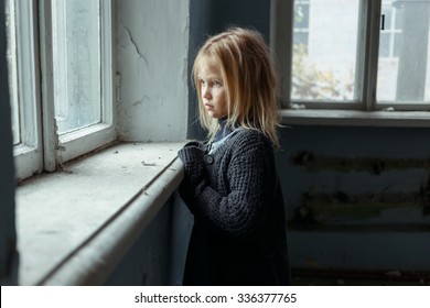 Hopeless life. Close up of depressed poor little girl standing near window and looking aside while feeling miserable