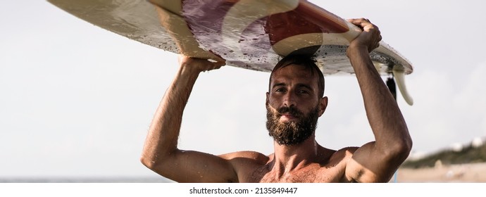 Horizontal banner or header with surfer holding his surfboard on the head - Hipster man standing on the beach and waiting big waves for surfing - Fit bearded man training with surfboard to sea