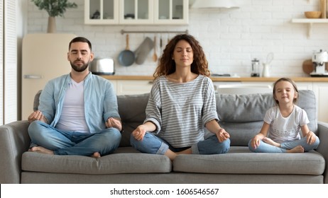 Horizontal banner serene couple and little daughter sitting on sofa in lotus position closed eyes do meditation breathing technique in living room, keep calm, healthy life habits and lifestyle concept - Shutterstock ID 1606546567