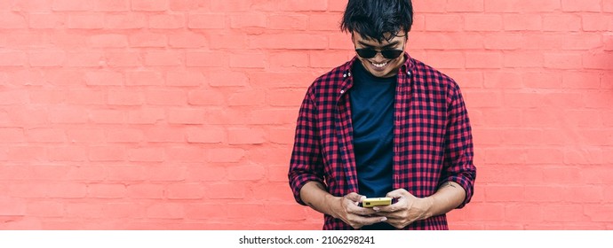 Horizontal banner with smiling  young guy using mobile phone outdoor against a red wall - Filipino social influencer having fun with new trends smartphone apps - Focus on face
