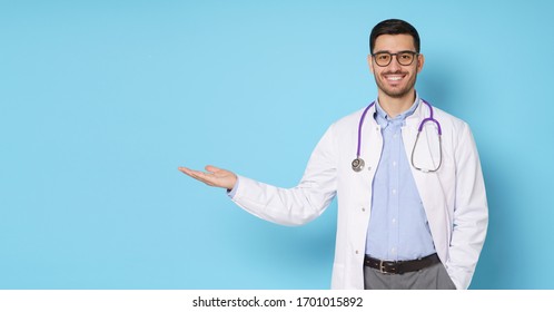 Horizontal banner of smiling young male doctor showing and presenting something with hand, isolated on blue background with copy space on the left side