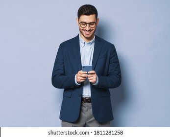 Horizontal Banner Of Young Business Man Wearing Glasses And Formal Clothes, Texting On Phone, Smiling Happily At Screen, Isolated On Blue Background