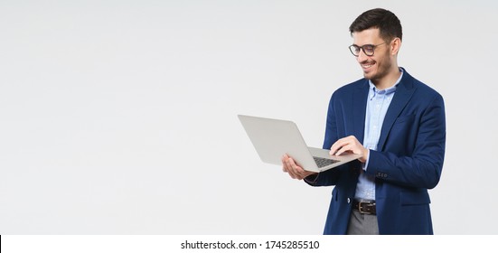 Horizontal banner of young handsome smiling business man holding laptop in hands, typing and browsing web pages, isolated on gray background with copy space