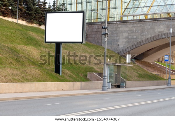 Horizontal billboard on a green lawn next to a\
pedestrian bridge and a bus\
stop.