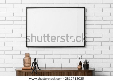 Horizontal black frame mockup on a white brick wall with a wooden cabinet and home decor items
