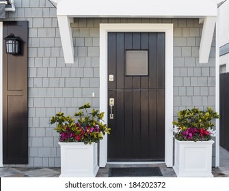 Horizontal Of Black Front Door To A Family Home; The Door Is Vertical Wood Boards, With A Window, Framed By Two Planters, Gray Shingles, And A Door Mat. Also Seen Is A Porch Light Fixture. 