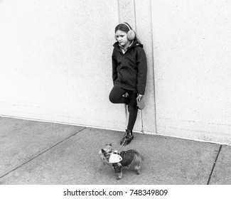 Horizontal Black And White Photo Of Forlorn-looking Little Girl In Fall Jacket And Ear Muffs Leaning With One Foot On Wall While Holding Tiny Yorkshire Terrier Dog On Leash