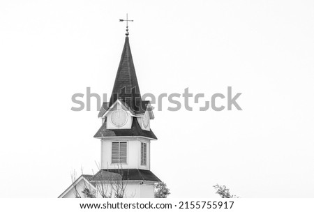 Horizontal black and white shot of top of European style church clock tower’s spire with clock in Roman numerals about to strike 9am