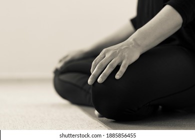 Horizontal black and white side detail of yogini on the floor with legs crossed in lotus pose. Woman indoors wearing black yoga pants with hands resting on knees. Home yoga practice in sepia tone