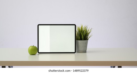 Horizontal blank screen tablet with tree pot and green apple on white wooden table and white wall background