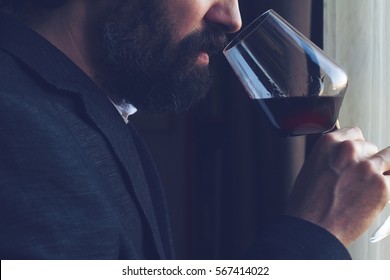 horizontal close up of a Caucasian man with beard, black suit and white shirt tasting a glass of red wine in front of a window