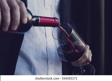 Horizontal close up of Caucasian man in black suit and white shirt pouring red wine into a tall glass in a bar - Powered by Shutterstock