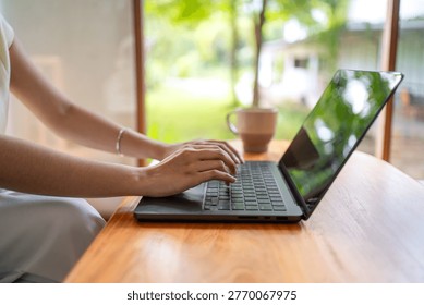 Horizontal close up of hands typing on laptop with coffee cup on wooden table in cozy home setting.