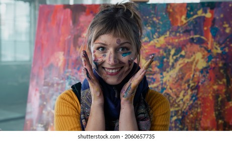 Horizontal Close Up Portrait Of A Caucasian Female Artist Covered In Paint, Looking Directly At The Camera, Smiling. Standing In Front Of Her Modern Abstract Art Piece In An Empty Warehouse Space. 