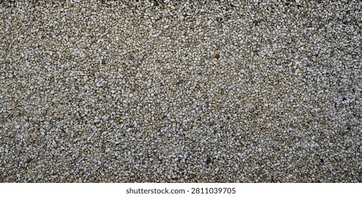 A horizontal close up of a wall surface with multi size gravel. The stones are various sizes, shapes, and colors including gray, tan, black, and reddish‑brown. The rocks are embedded in a cement mixture that adheres them to the wall. The stones are porous, some are broken, and the gravel is rough and uneven.
