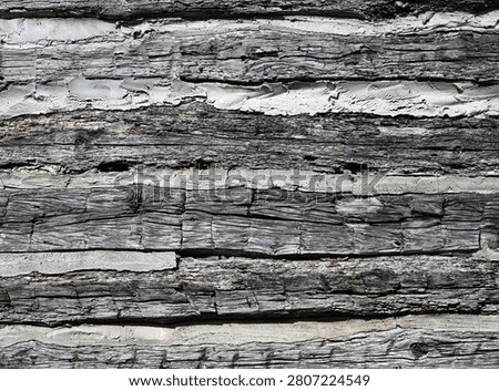 A horizontal close up of weathered log cabin logs with flat dried cracked and splintered wood showing strong grain, knots, and cut marks. Thick porous chinking fills the gaps between the logs and appears cracked, uneven, and missing in some areas.