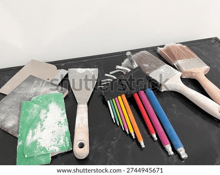 Horizontal close-up of a black table with slightly paint-stained brushes, colorful hand tools, grout tools, and a spatula, showing equipment used for home renovation and wall repair projects.