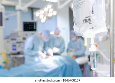 A horizontal closeup of an IV bag hanging in the foreground in selective focus with a team surgeons and nurses working in the background - Powered by Shutterstock