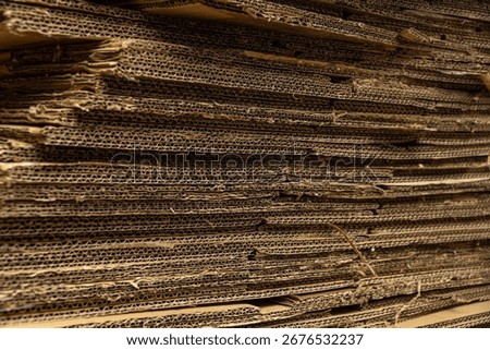 A horizontal close-up of a large stack of cardboard, showing the rough, corrugated texture and compressed layers.