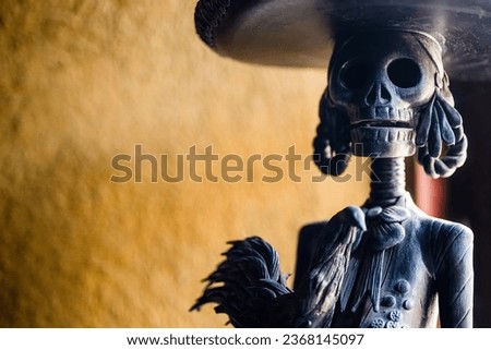 Horizontal close-up photo of a high-contrast black clay Catrina figure, dressed as a charro, holding a fighting rooster, set against a vibrant orange background.