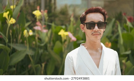 A horizontal close-up portrait shot of a non-binary gay man of Isan, Thai descent standing outdoors in bright daylight. The model has short dark hair with distinctive deep reddish-brown highlights, a mustache, black earrings on both earlobes, and is wearing an off-white or cream-colored wrap blazer jacket with a deep v-neck. Positioned on the right side of the frame, the individual is wearing large square black sunglasses and is looking directly forward toward the camera lens with a calm, neutral facial expression. Behind the subject on the left is a vibrant bed of large-leaf green tropical canna lily plants featuring colorful yellow and pink blossoms. In the background, a modern building facade with large glass windows reflects the bright ambient outdoor daylight. Shot with natural, soft diffused sunlight highlighting the model's authentic, unretouched skin texture and the woven linen-like pattern of the garment. The composition features an off-center placement with a very shallow depth of field, keeping the individual sharply in focus while smoothly blurring the botanical background on the left.
