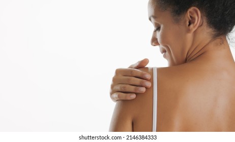 Horizontal Close-up Of Slim African American Woman Strokes Her Shoulder Standing With Her Back On White Background | Skin Moisturizing And Body Care Concept