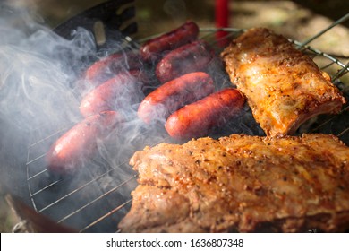 Horizontal Close-up Of A Smoky Father's Day Barbecue, With Sausages And Pork Cuts Being Grilled Under A Contrasty Chiaroscuro Light. Outdoor Meal 