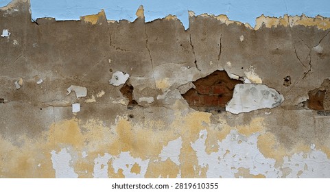 A horizontal close-up view of a bedraggled multi-layer wall surface with weatherworn paint, concrete, and red brick with mortar. The light blue, white, and yellow paint is flaking, cracking, chipping, and peeling off the concrete layer. The concrete is the largest visible area and is porous, rough, uneven, cracked, and full of holes that reveal small areas of worn terra cotta cement and red bricks with weatherworn rough, uneven faces and mortar joints wearing away.