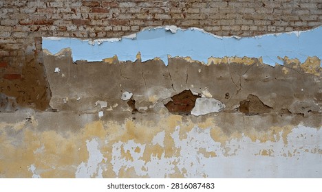 A horizontal close-up view of a decaying multi-layer wall surface with weatherworn paint, concrete, terra cotta cement, and red brick with mortar. The light blue, white, and yellow paint is flaking, cracking, chipping, and peeling off the concrete layer. The concrete is porous, rough, uneven, cracked, and breaking away to reveal small areas of worn terra cotta cement and a large exposed red brick layer with rough uneven bricks covered in mortar and white paint in places.