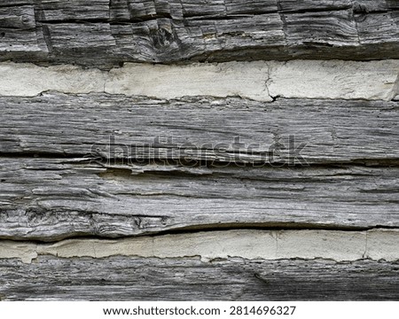 A horizontal close-up view of a weathered log cabin wall with flat dried splintered and cracked logs showing visible wood grain, knots, and cut marks. The chinking fills the gaps between the logs and is porous, cracked, and uneven.