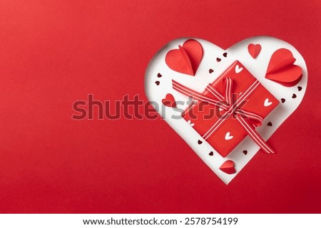 A horizontal composition featuring a red heart-shaped cutout with a gift box tied with a striped ribbon and surrounded by paper heart decorations.