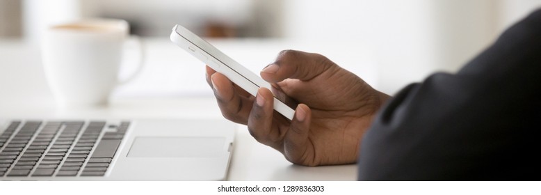Horizontal concept photo african businessman sitting at desk near pc close up hand hold use smartphone app communicate online browse internet, banner for website header design with copy space for text