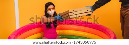 horizontal crop of delivery man giving pizza boxes to woman in swimsuit, medical mask and sunglasses on yellow