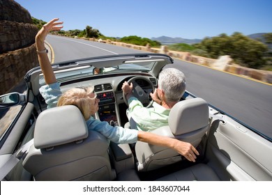 Horizontal Elevated Rear View Of A Joyous Couple Driving A Convertible With The Woman Spreading Arms On An Highway Besides Coast At A High Speed With Motion Blur On A Sunny Day.