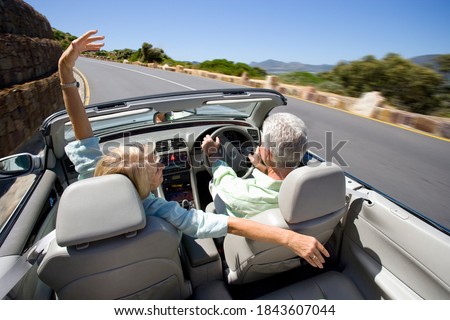Horizontal elevated rear view of a joyous couple driving a convertible with the woman spreading arms on an highway besides coast at a high speed with motion blur on a sunny day.
